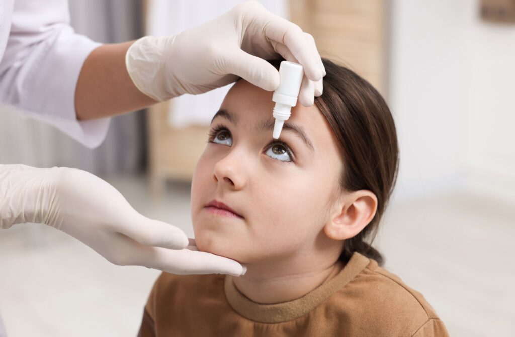 An optometrist holds a young child’s chin to keep their head steady while applying eye drops to relief allergy symptoms.