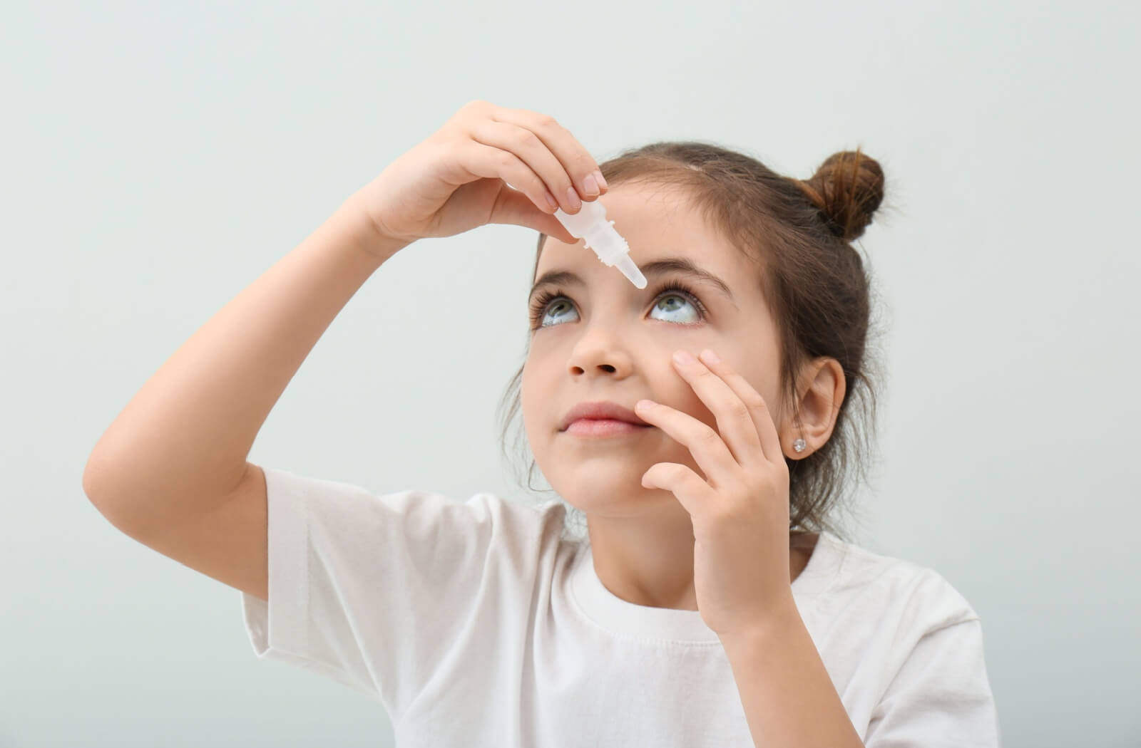 Against a plain background, a young child in white holds their eyelid down to make it easier to apply eye drops on their own.