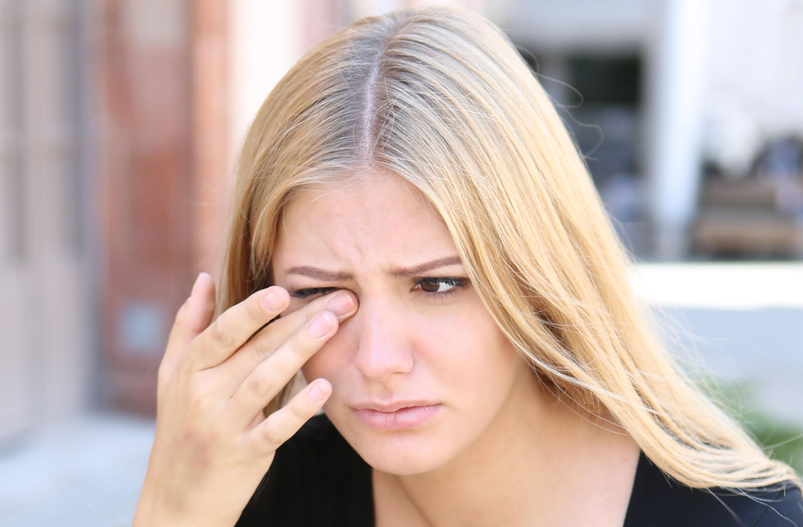 A young adult rubbing their right eye outdoors in frustration due to a stuck contact lens still in their eye.