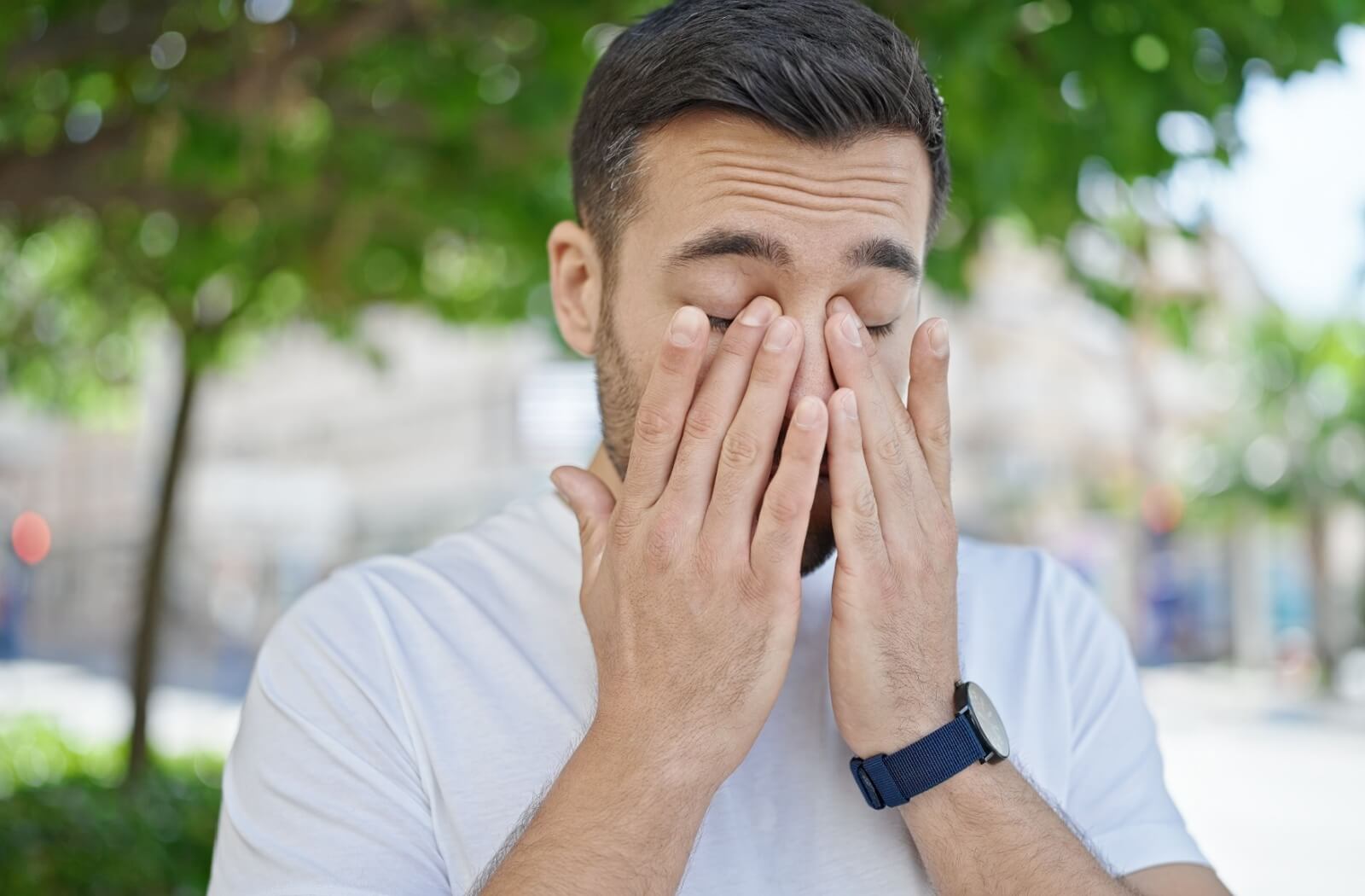 A person standing outside rubs their eyes due to the pollen that's causing their allergies to trigger.