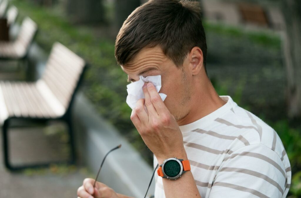 Man wiping his irritated eye with a tissue while holding his eyeglasses outdoors.